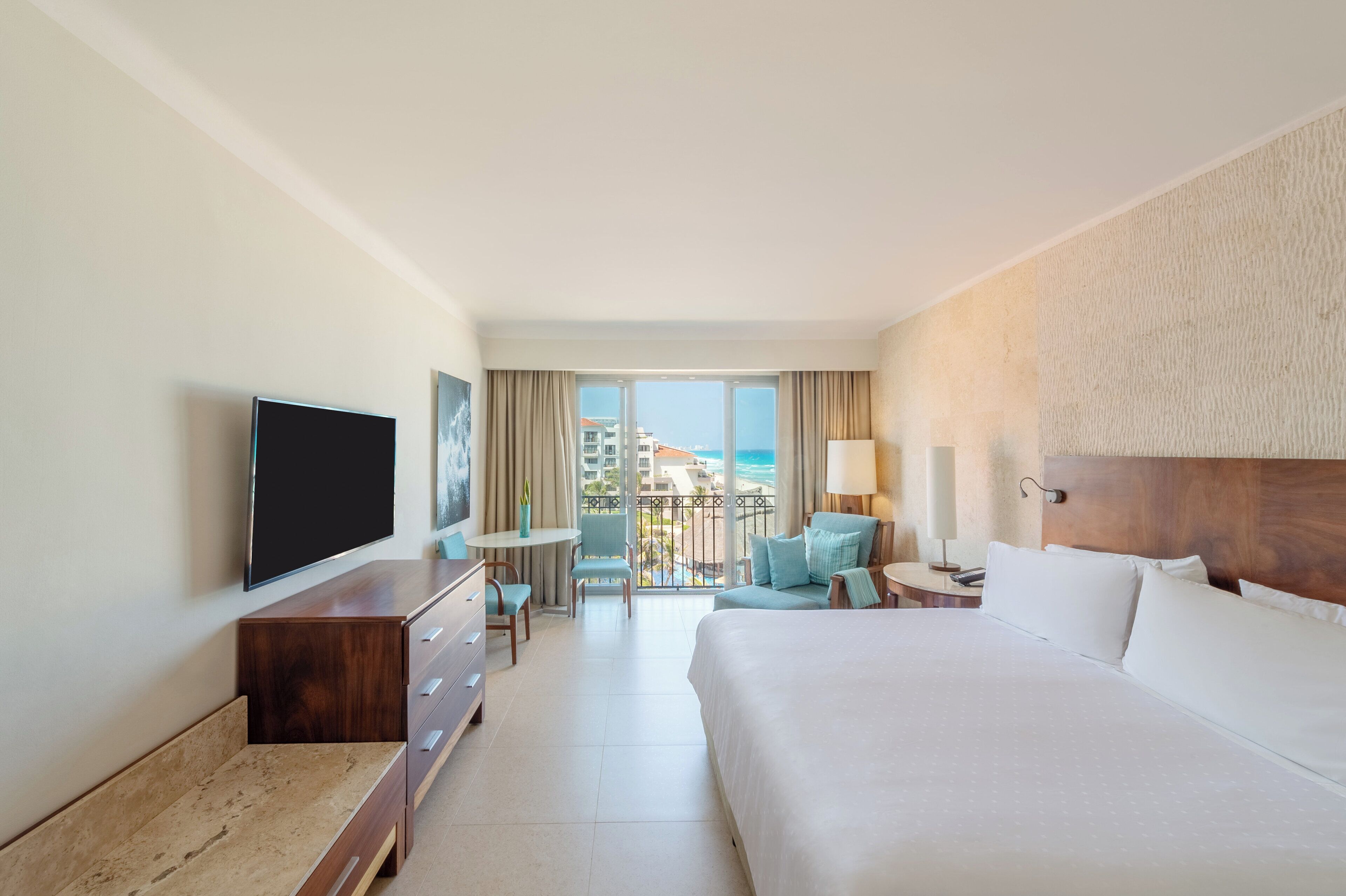 A hotel room with a large bed, a flat-screen TV, a desk with a chair, and a balcony with a view of the ocean.