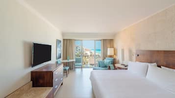 A hotel room with a large bed, a flat-screen TV, a desk with a chair, and a balcony with a view of the ocean.