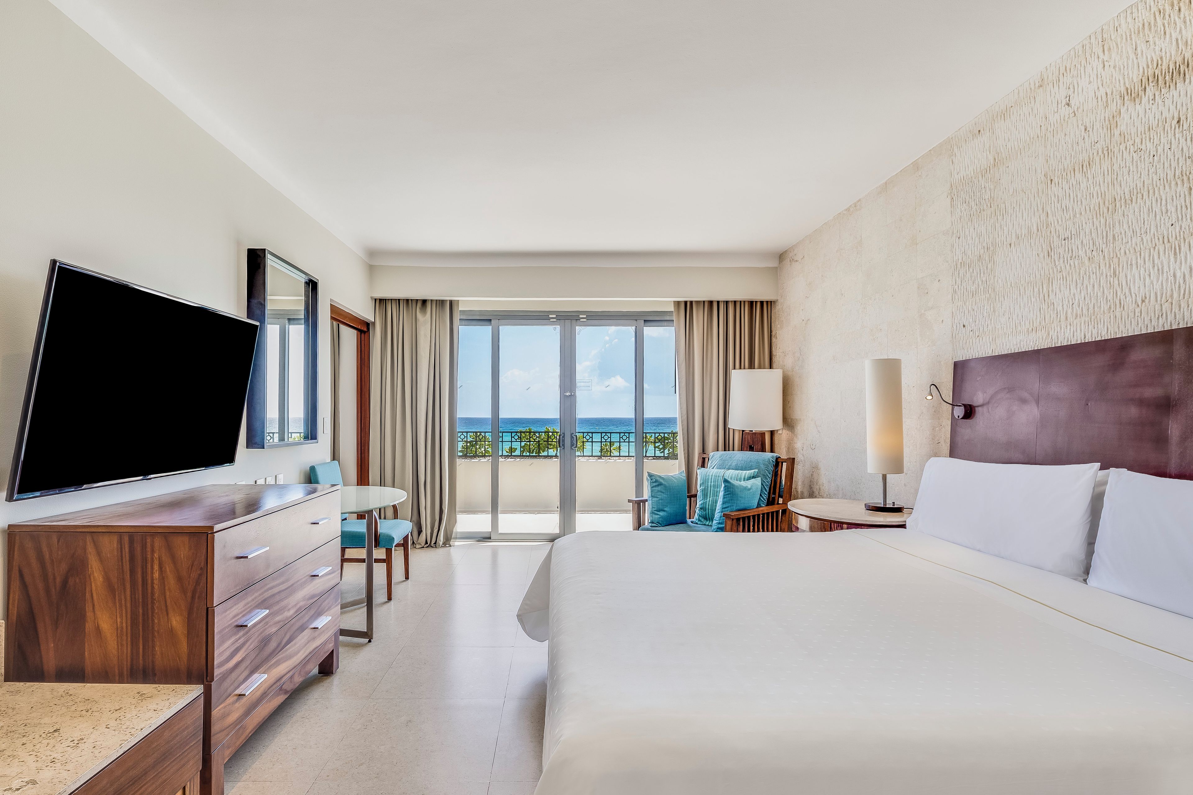 A hotel room with a bed, a TV, a desk, a chair, a lamp, and a view of the ocean.