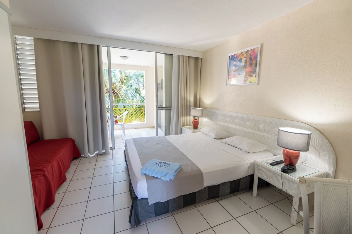 Quarto de hotel com cama, mesa de cabeceira, uma cadeira vermelha, varanda com cortinas e um quadro na parede.