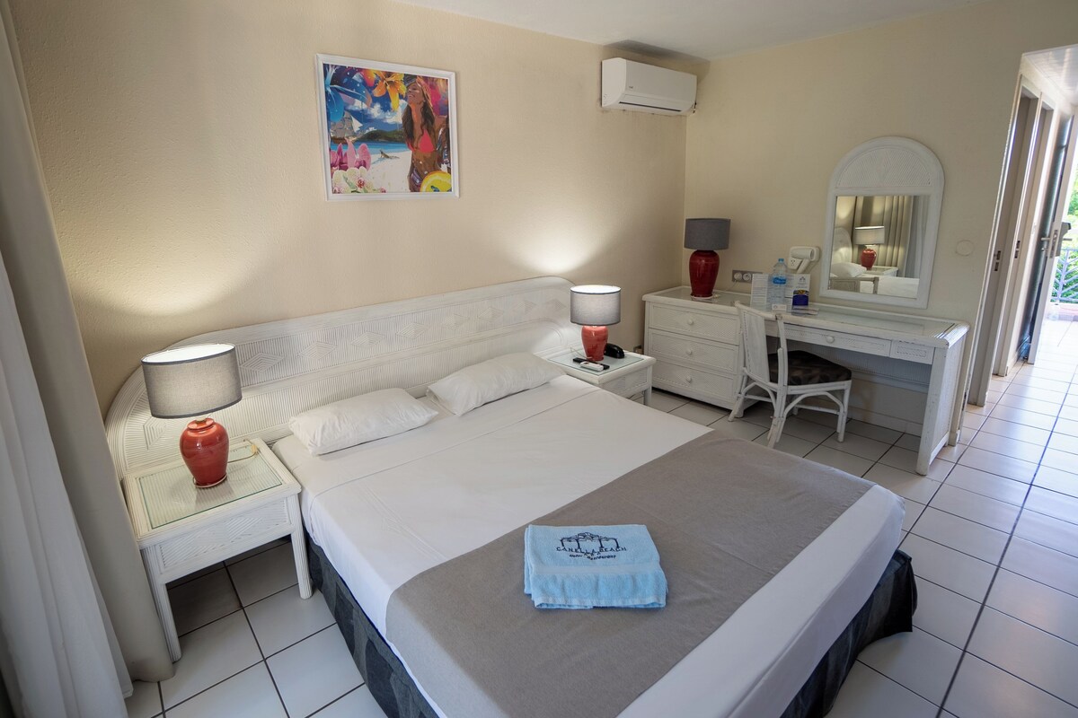 Quarto de hotel com uma cama grande, mesas de cabeceira, uma escrivaninha com cadeira, um espelho e um quadro na parede.