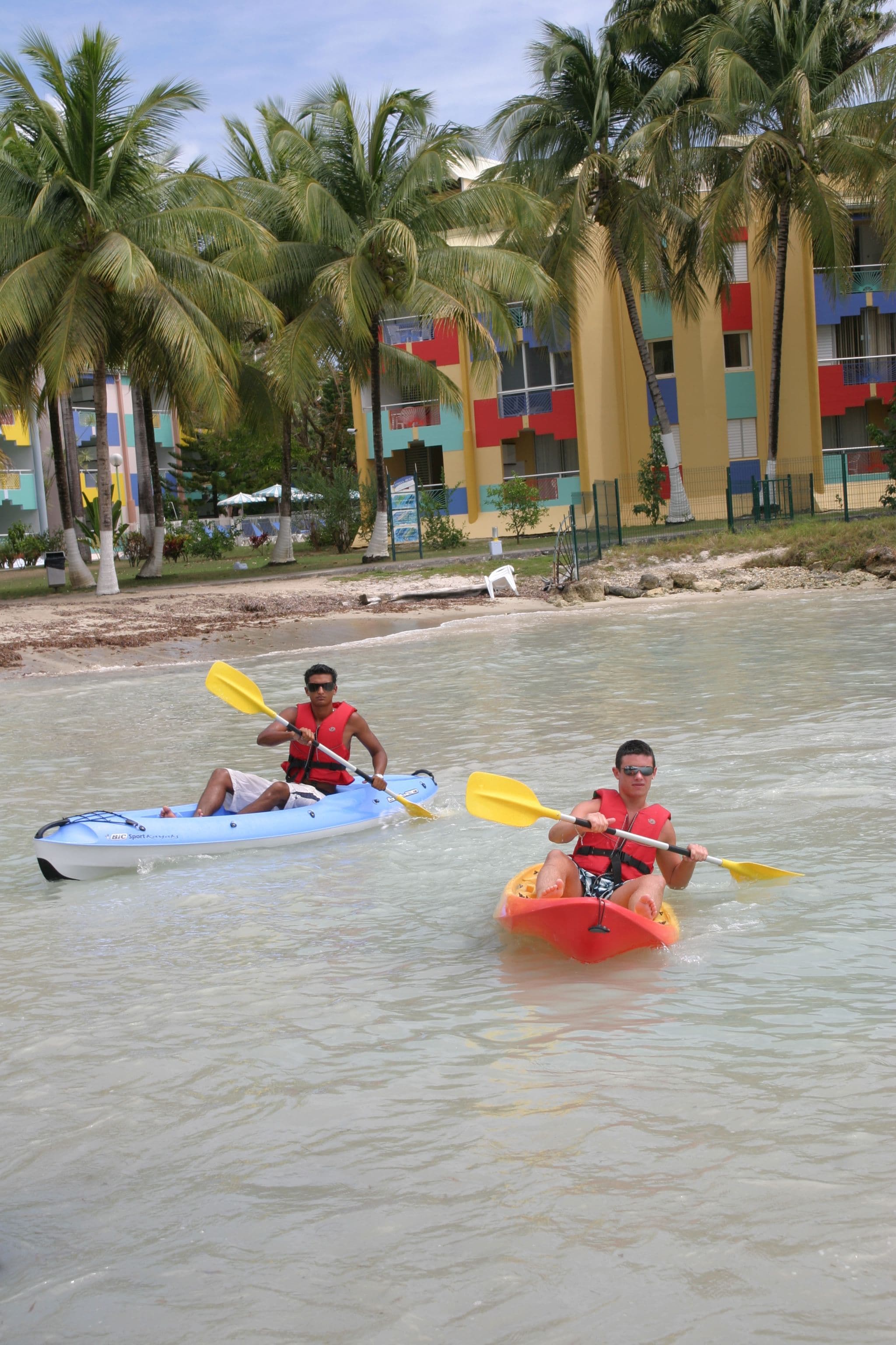 Two people kayaking on a calm river with palm trees and colorful buildings in the background.