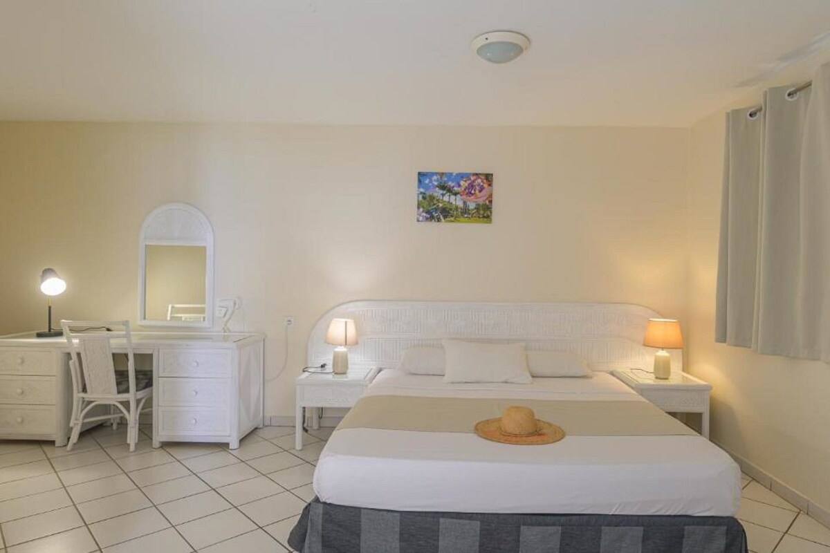 Quarto de hotel com cama, escrivaninha com cadeira, cômoda e um quadro na parede.