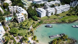 An aerial view of a coastal resort with multiple buildings, a sandy beach, palm trees, and a calm bay with boats. - Canella Beach Hotel (Le Gosier)