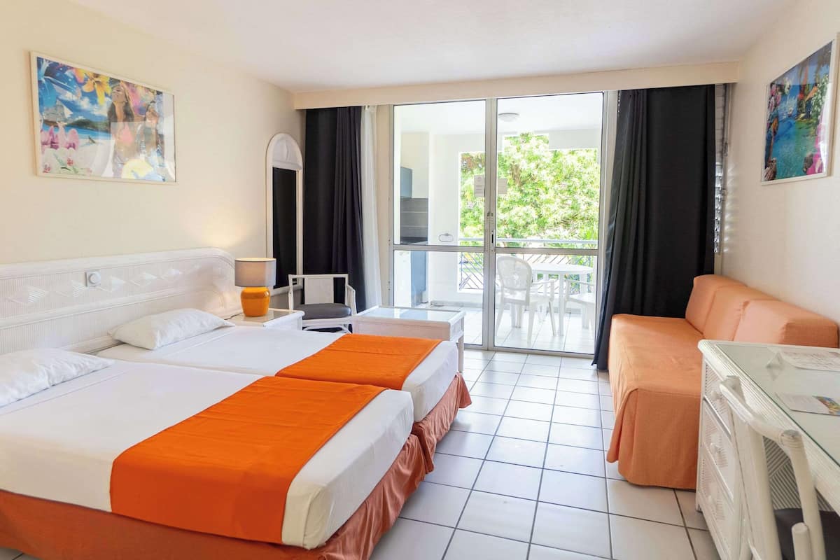 A hotel room with two beds, a sofa, a table, and a balcony with a view of greenery.