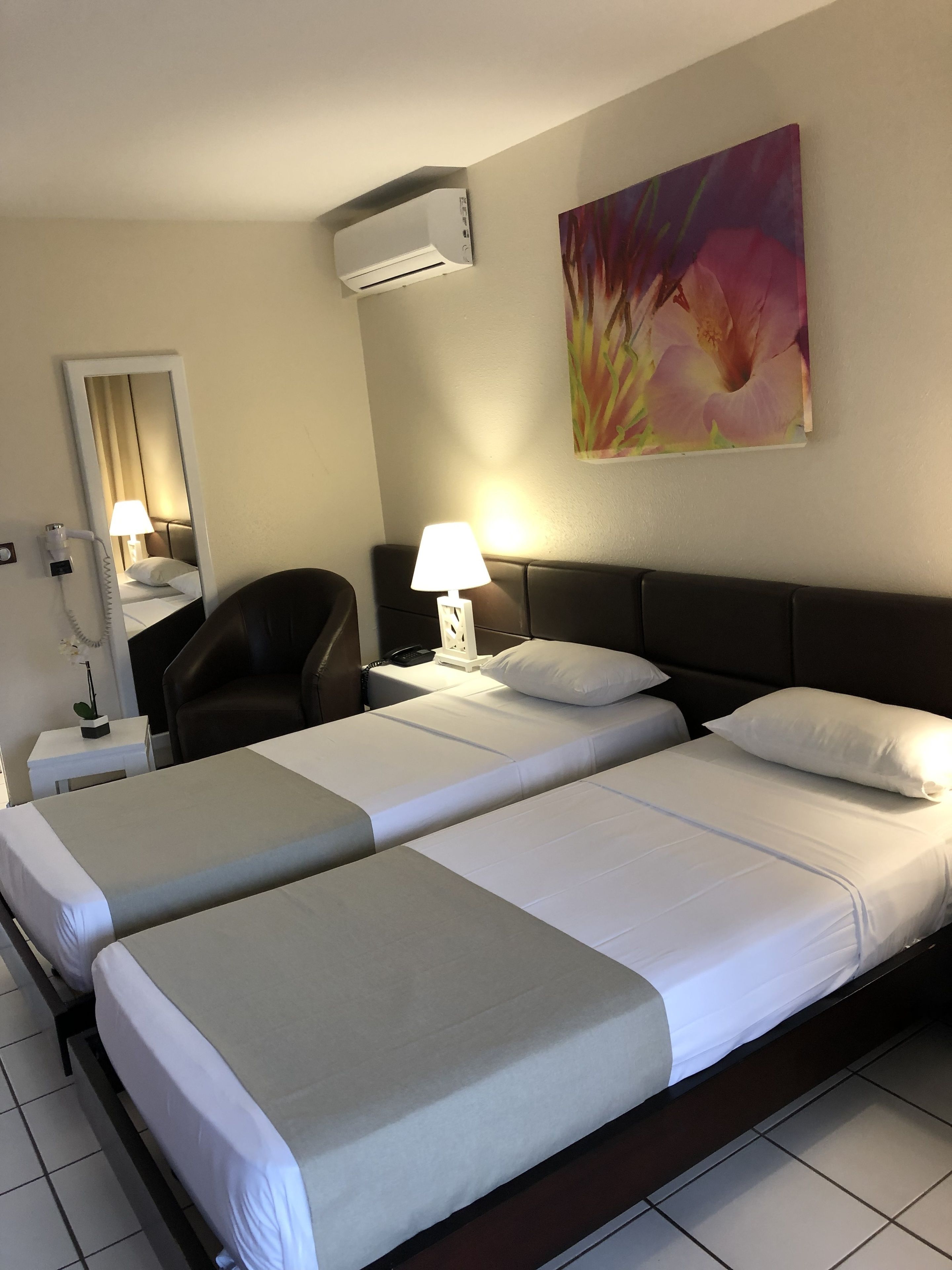 A hotel room with two beds, a desk, a chair, a lamp, an air conditioning unit, and a painting on the wall.