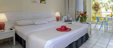 Ein Hotelzimmer mit einem großen Bett, einem Nachttisch, einem Sessel, einem Tisch und Blick auf Palmen durch das Fenster.