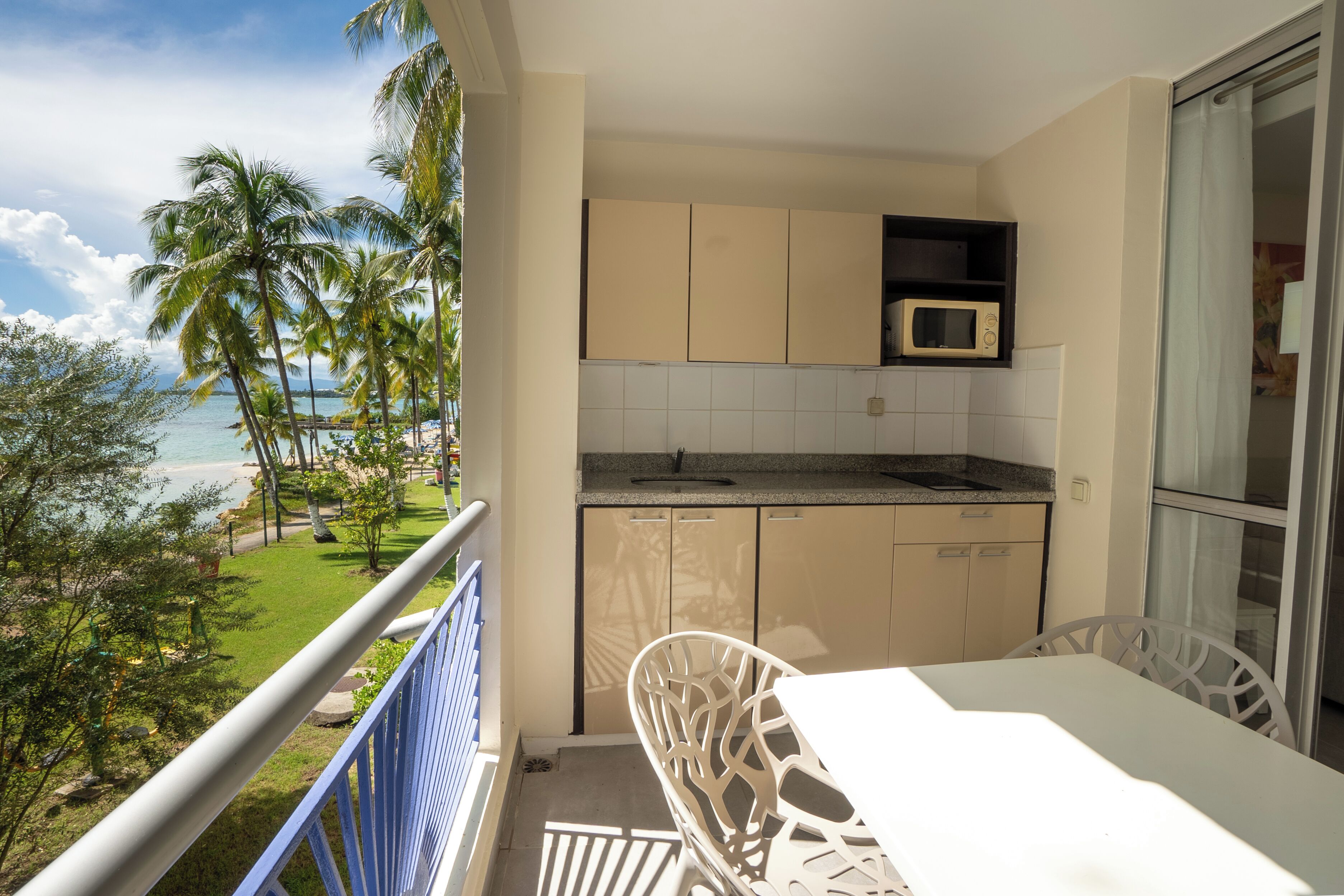 A balcony with a view of the beach, a white table, and a kitchen area with cabinets and a microwave.