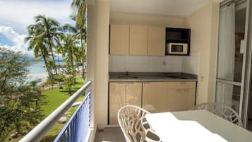 A balcony with a view of the beach, a white table, and a kitchen area with cabinets and a microwave.