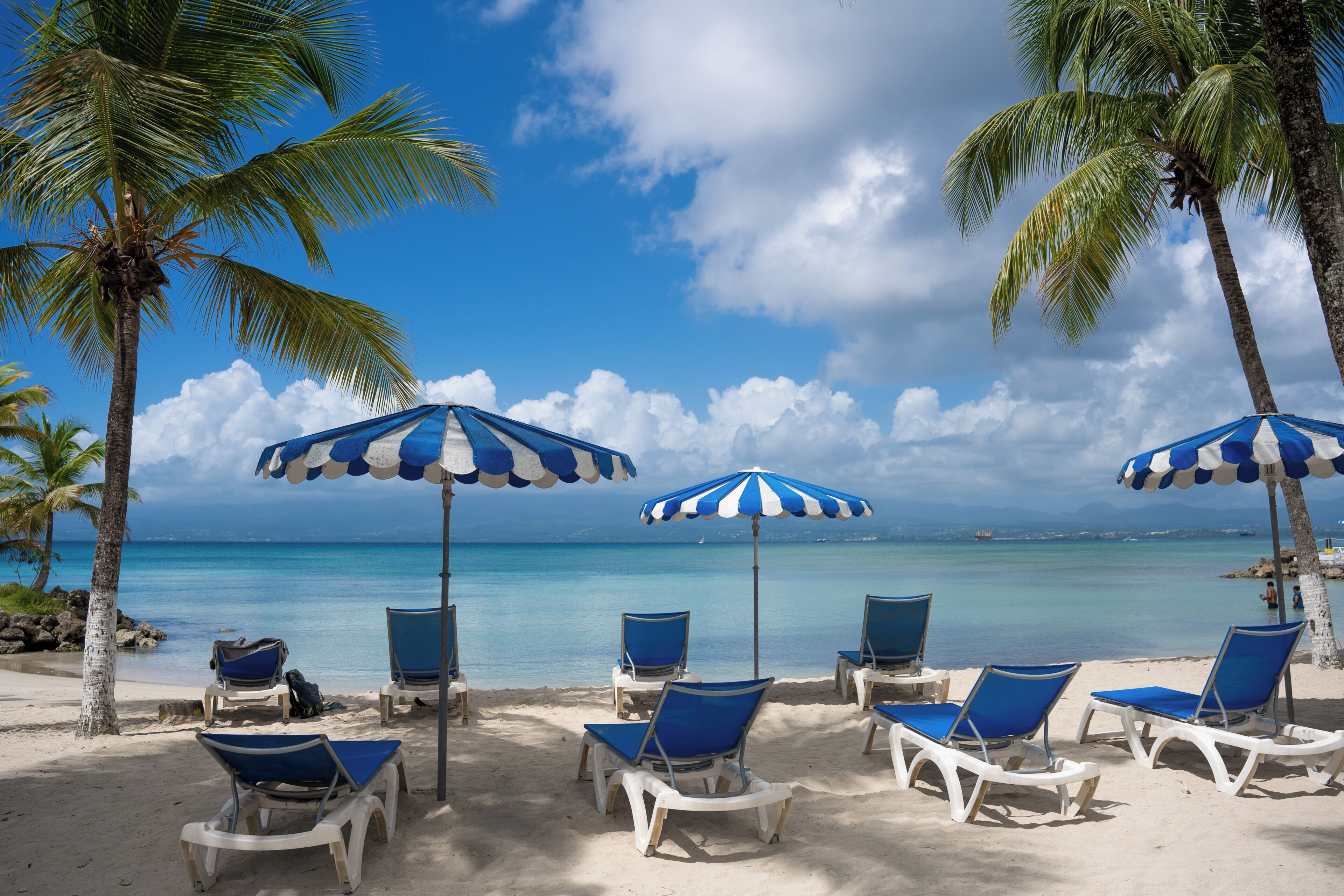 Une plage avec des chaises longues blanches et des parasols bleus, des palmiers et une mer d’un bleu limpide.