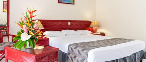 A hotel room with a bed, a nightstand, a lamp, a red chair, a table, and a decorative plant.