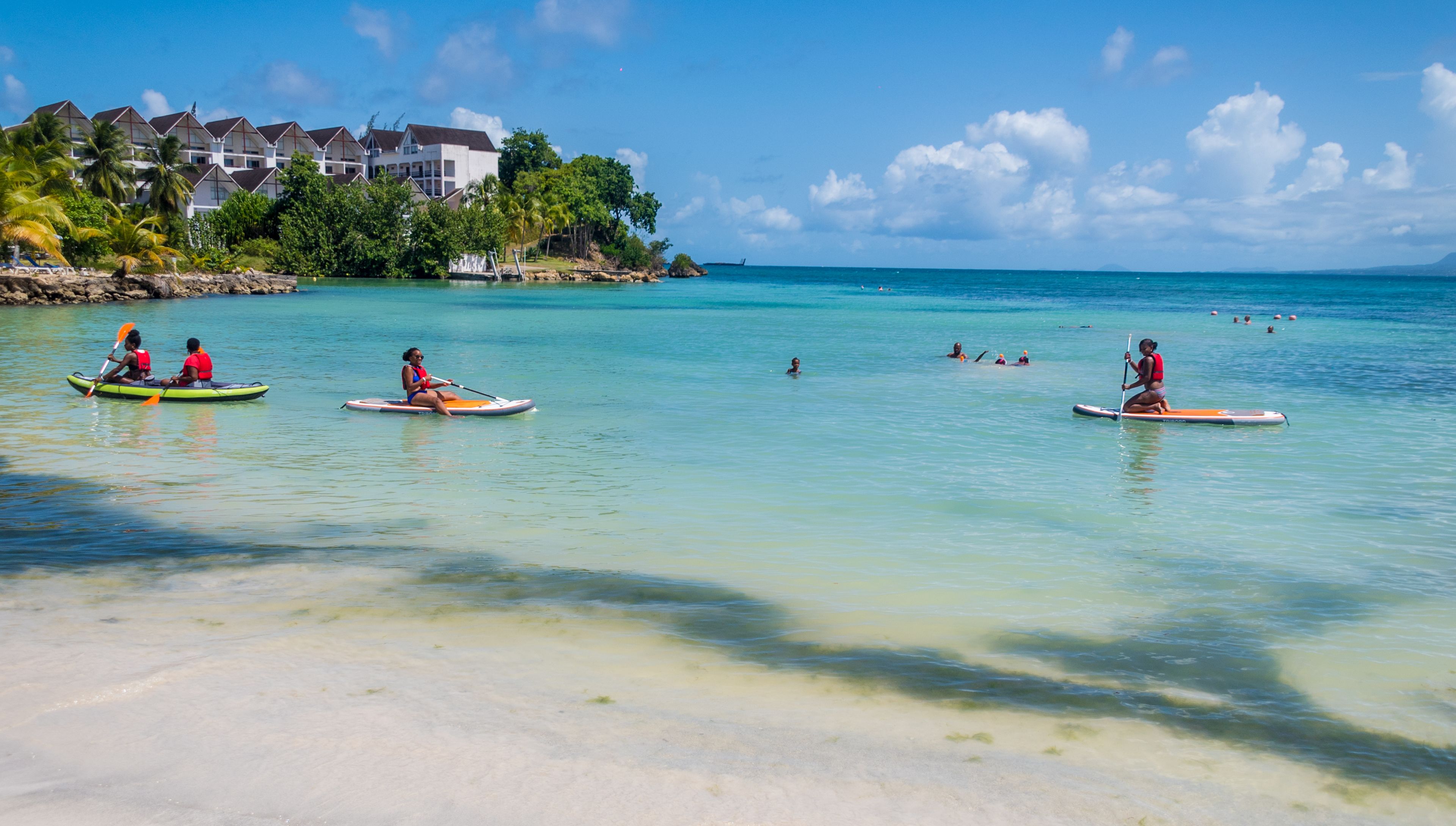Une plage à l’eau turquoise et limpide, un rivage sablonneux et un complexe hôtelier composé de plusieurs bâtiments en arrière-plan.