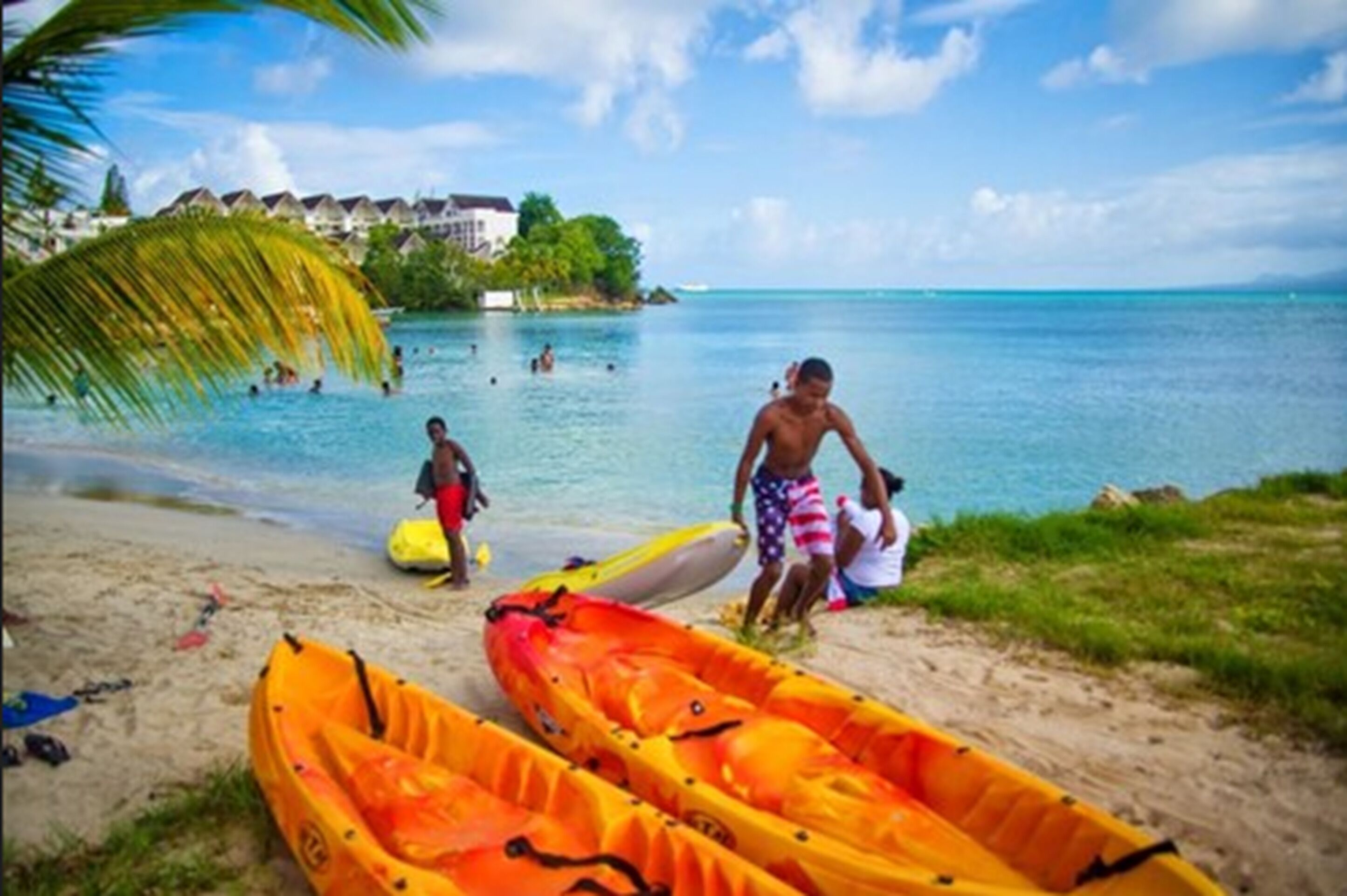 A beach with kayaks, a clear blue sea, and a resort in the background.