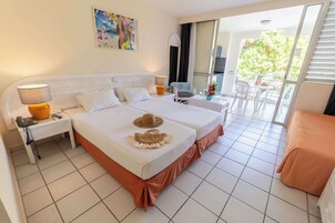 Habitación de hotel con dos camas, una mesita con lámpara, una silla y un balcón con vistas a árboles.