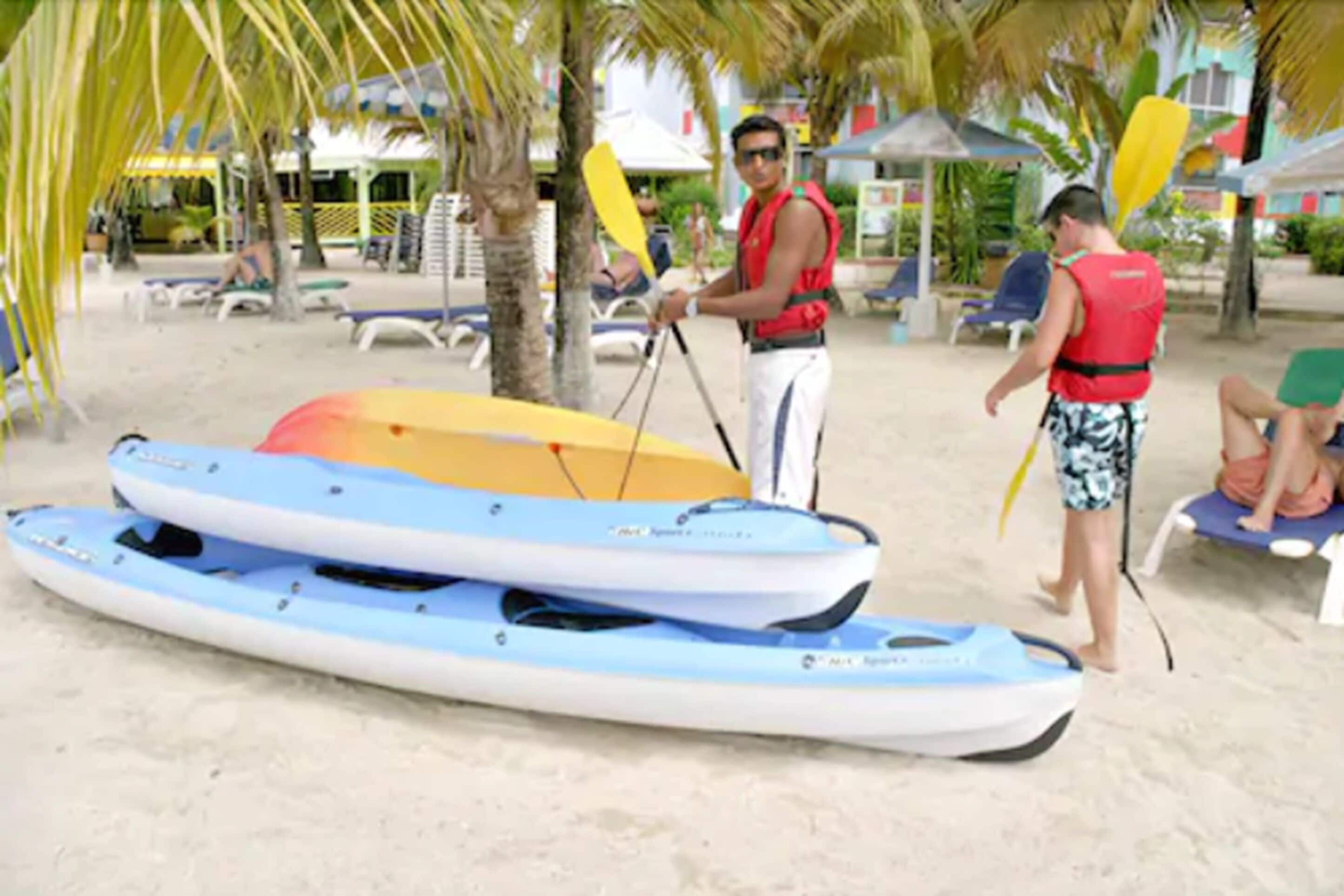 Una playa con kayaks, tablas de remo, palmeras y sillas de playa.