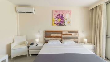 Habitación de hotel con cama, una silla, mesita de noche, aire acondicionado, un cuadro y ventana con cortinas.