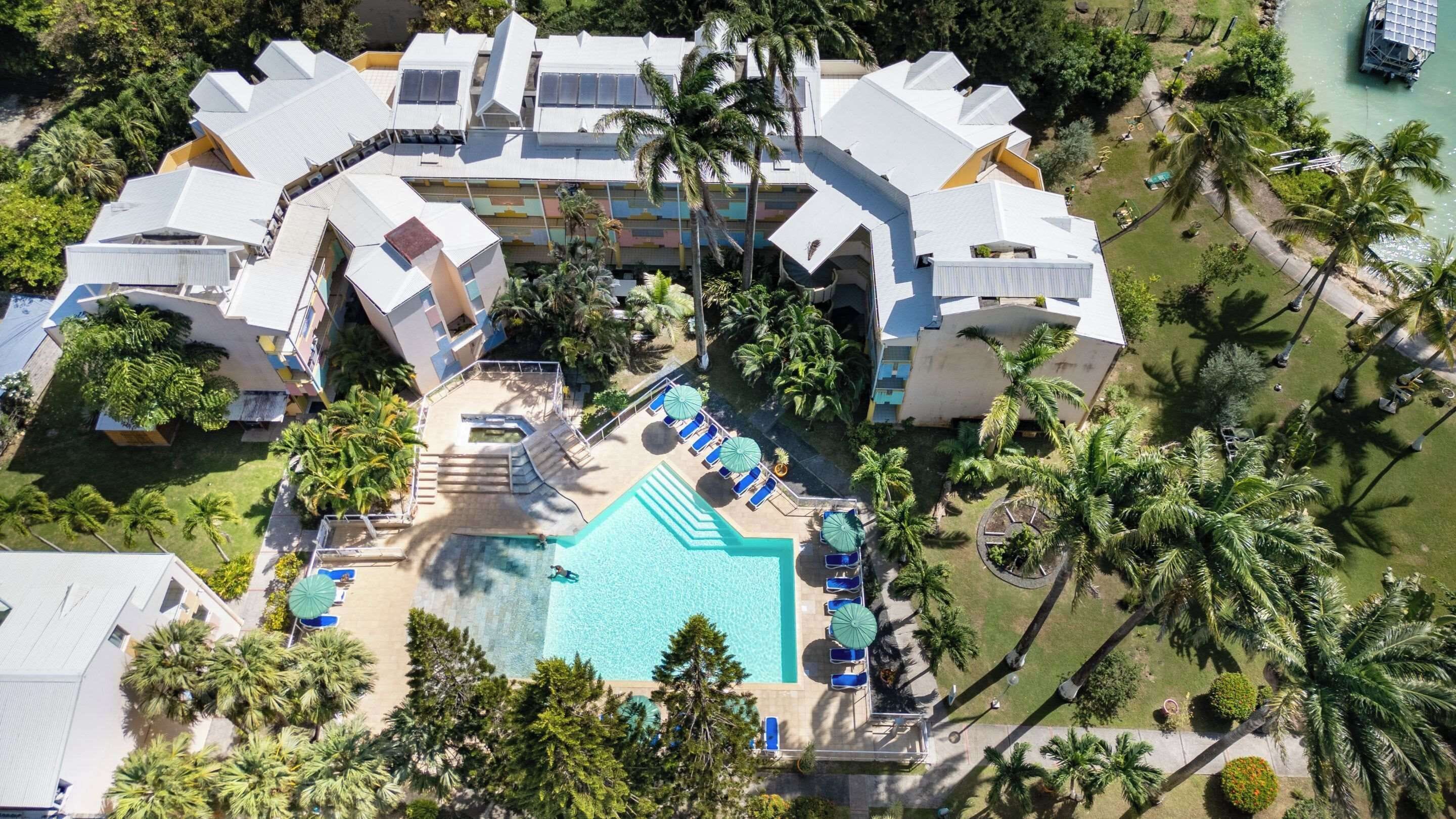 Vista aérea de un complejo turístico con piscina central rodeada de sillas de descanso y sombrillas, palmeras y una masa de agua.