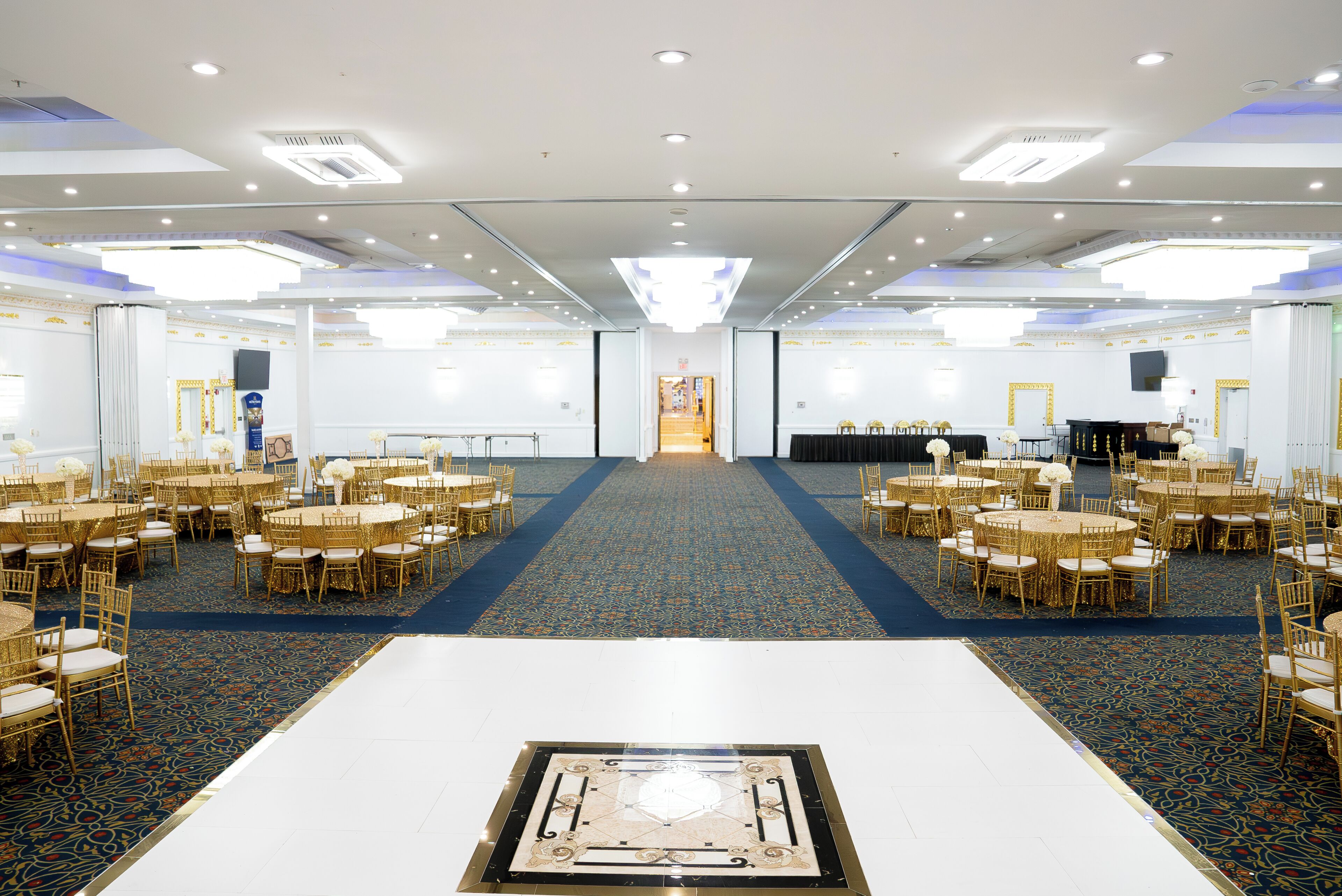 Een ruime congreszaal met meerdere tafels en stoelen, een kamerbreed tapijt met dessin, en een centraal decoratief element.