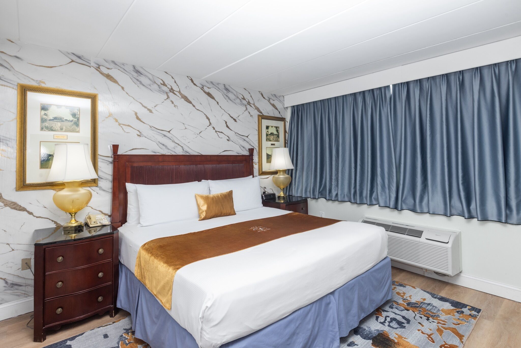 A hotel room with a marble-patterned wall, a bed with a wooden headboard, a blue curtain, and a nightstand with a lamp.