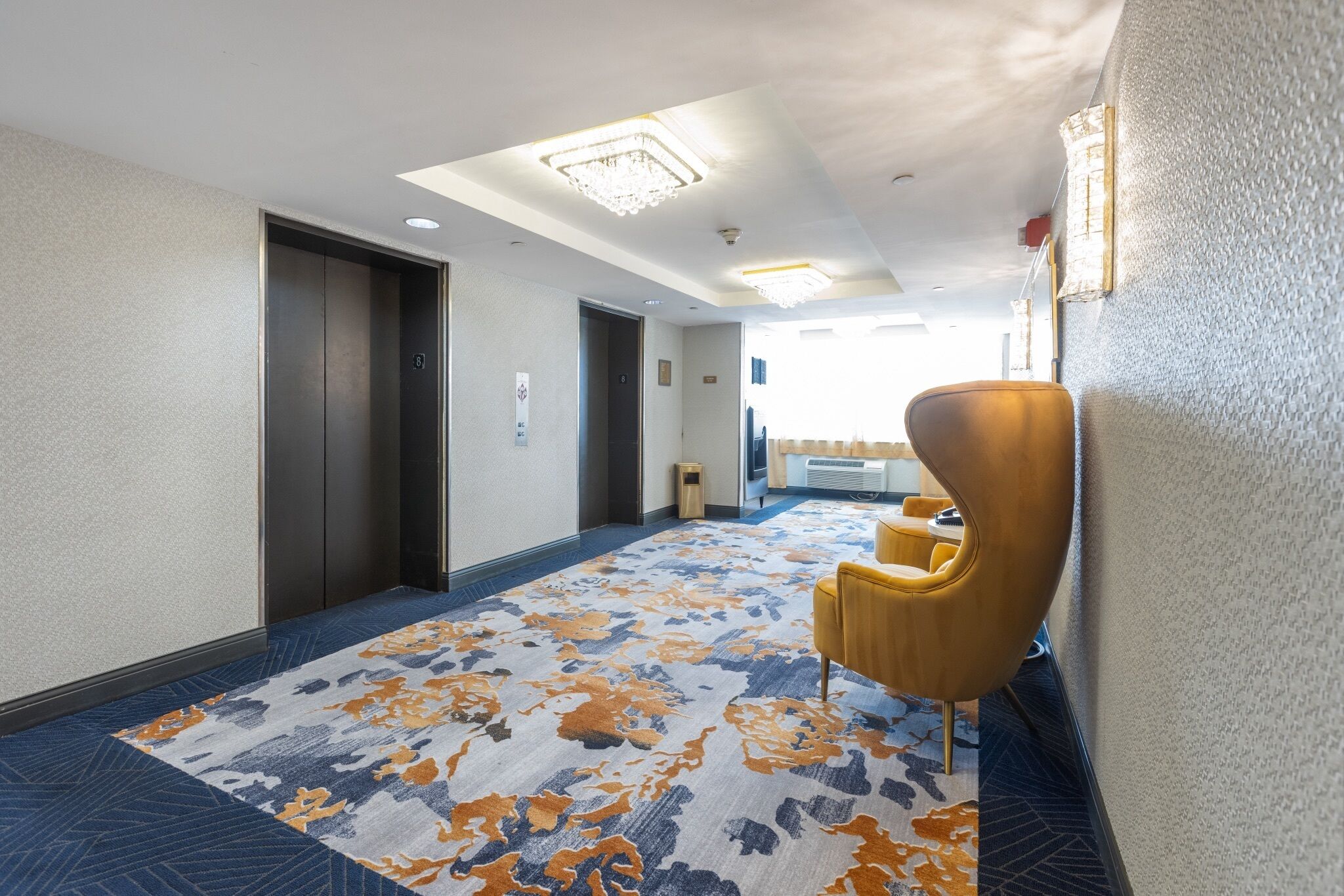 Een hotelgang met een kamerbreed tapijt, een gele fauteuil en een deur met een lamp erboven.