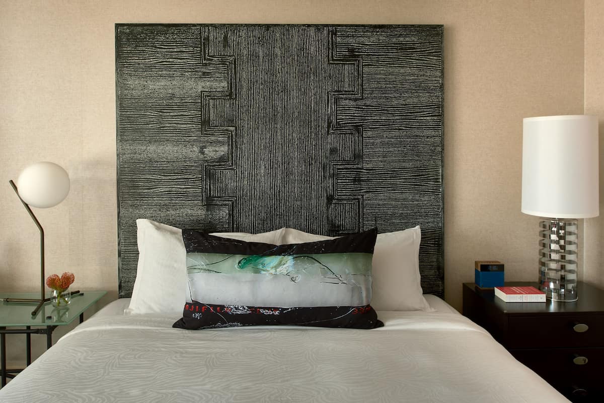 Una habitación de hotel con cama, mesita de noche, lámpara y una obra de arte en la pared.