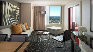 A hotel room with a sofa, a dining table, a chair, a small table, a floor lamp, a view of the city, and a window with curtains.