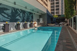 Una piscina con solárium empedrado, una pared con un mural de un árbol, y un edificio de varios pisos al fondo.