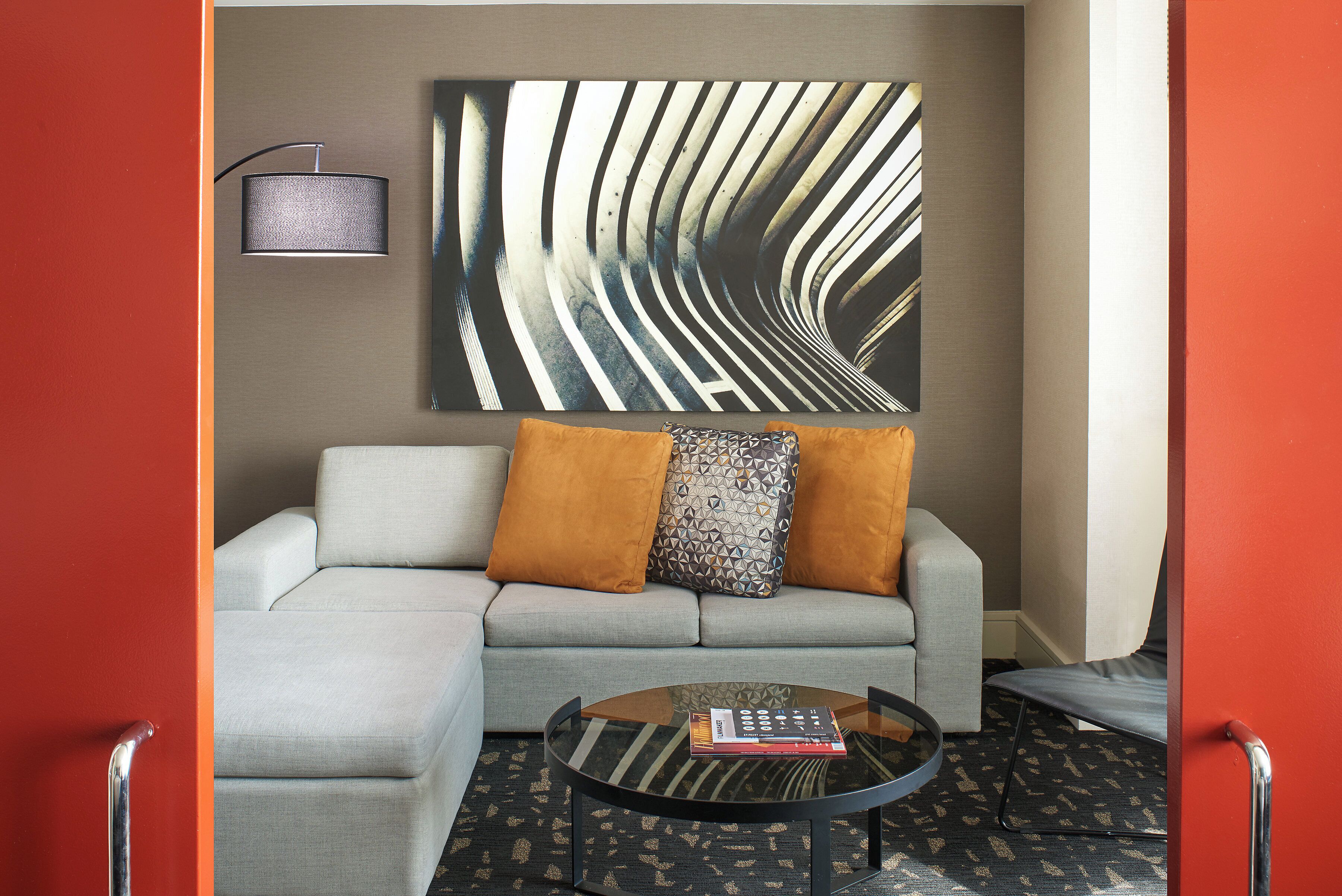 A modern living room with a grey sectional sofa, two orange cushions, a patterned throw, a glass coffee table with a book, and a wall-mounted lamp.