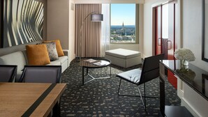 Een moderne hotelkamer met een bank, voetenbankje, eettafel en uitzicht op de stad door grote ramen.