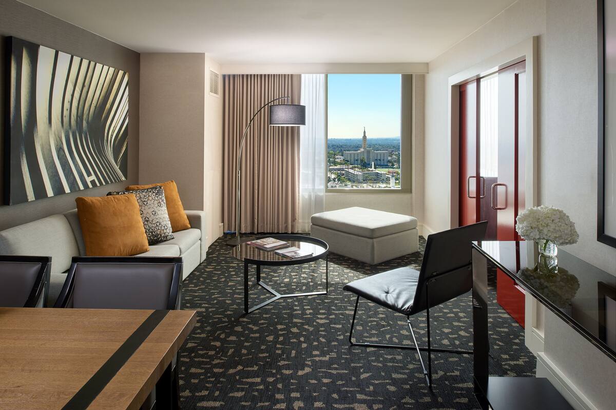 Una habitación de hotel moderna con sofá, reposapiés, mesa de comedor y vistas a la ciudad a través de amplias ventanas.