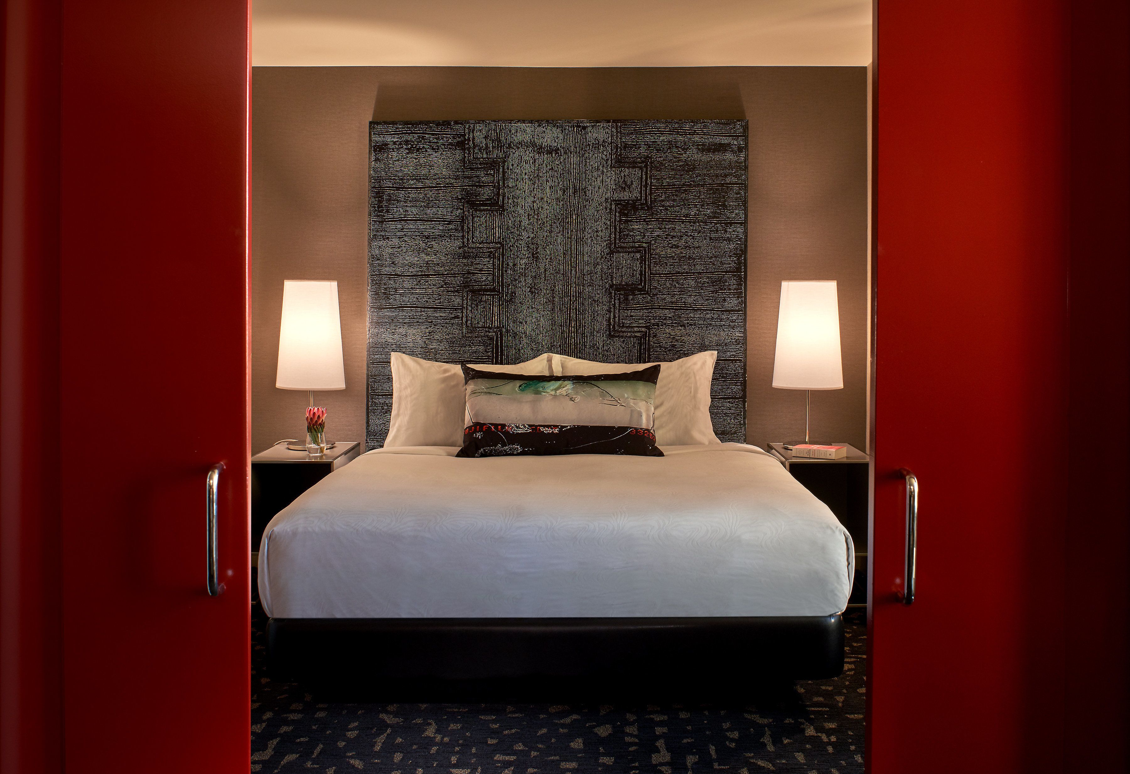 A hotel room with a large bed, two bedside lamps, a decorative headboard, and a red door.