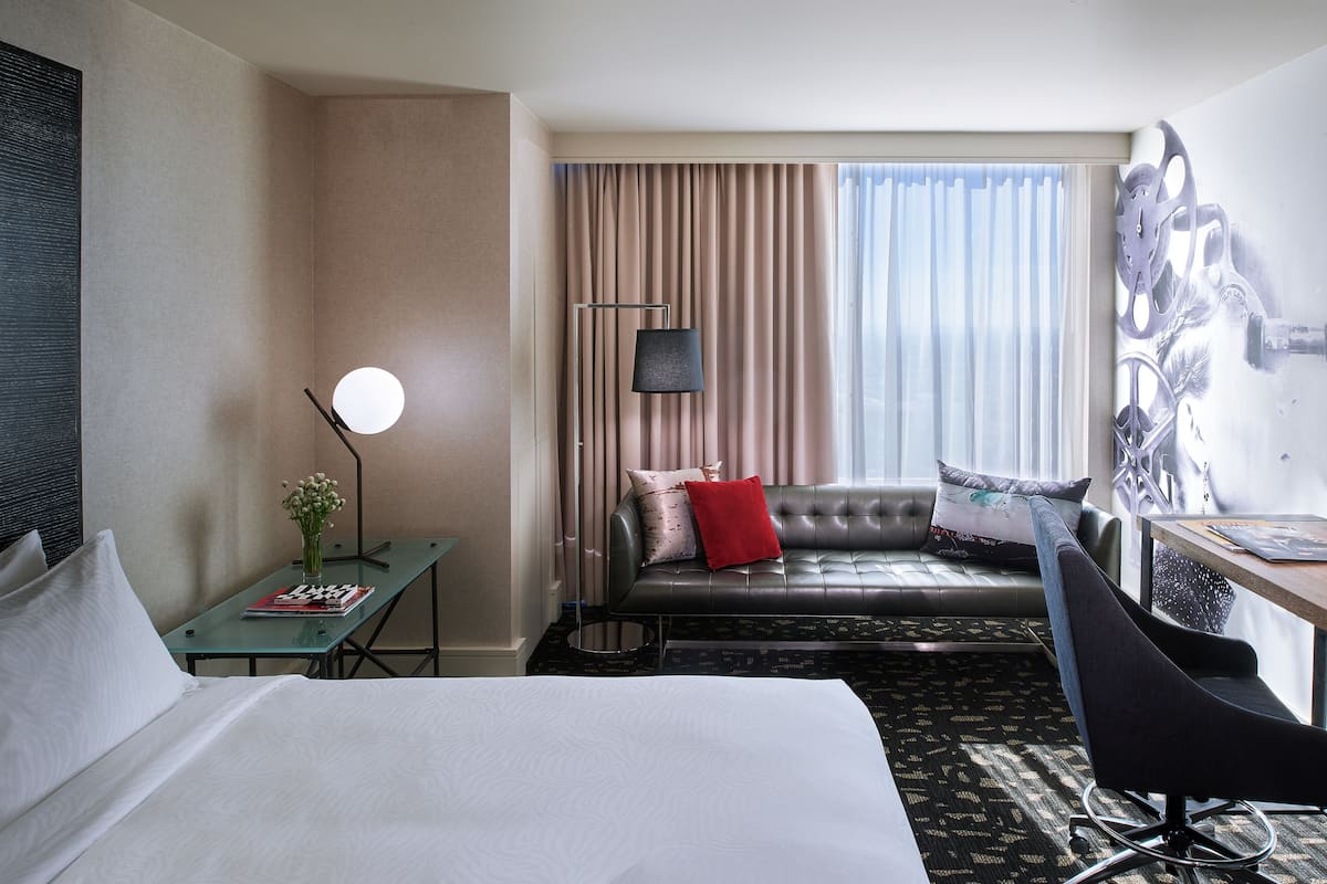 Habitación de hotel con cama, sofá, escritorio, silla, lámpara y ventana con cortinas.