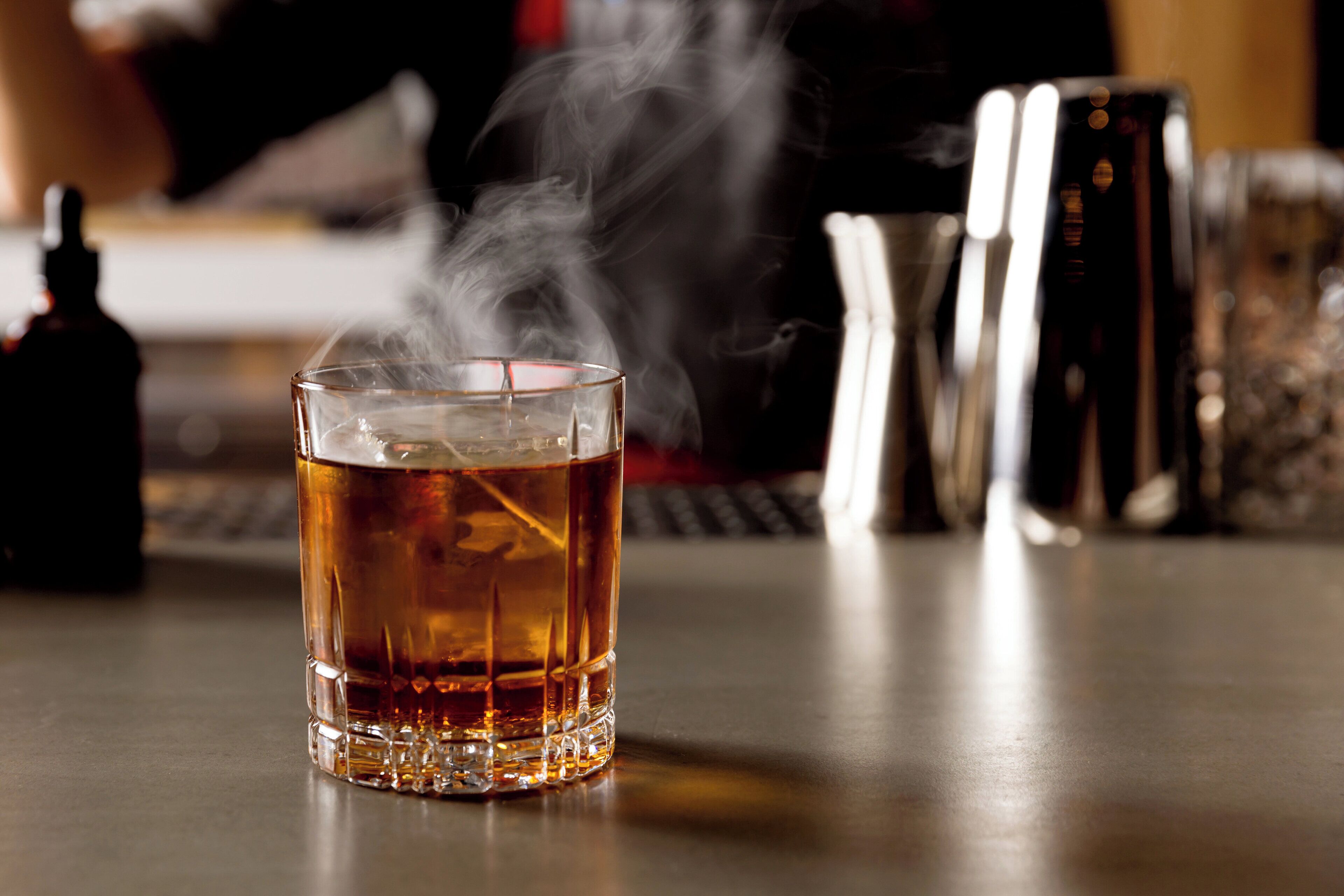 Un vaso de líquido ámbar con cubitos de hielo, vapor saliendo de él, y un bar con una botella y un coctelero al fondo.