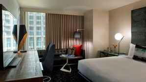 Ein Hotelzimmer mit einem Bett, einem Schreibtisch mit Laptop, einem Stuhl, einem Sofa, einem Fernseher und einem Fenster mit Vorhängen.