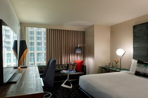 Et hotelværelse med seng, skrivebord med computer, stol, sofa, fjernsyn og et vindue med gardiner.