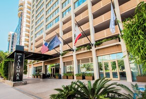 Entrada de un hotel con un cartel que dice 'PALOMAR HOTEL' y banderas, incluyendo la bandera estadounidense.