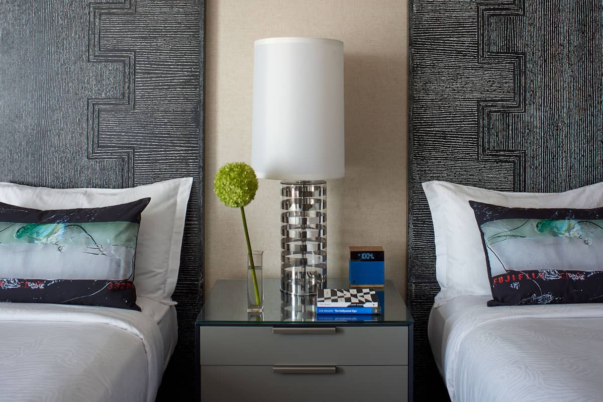 Habitación de hotel con dos camas, una mesita de noche con lámpara, un reloj pequeño y un jarrón decorativo.