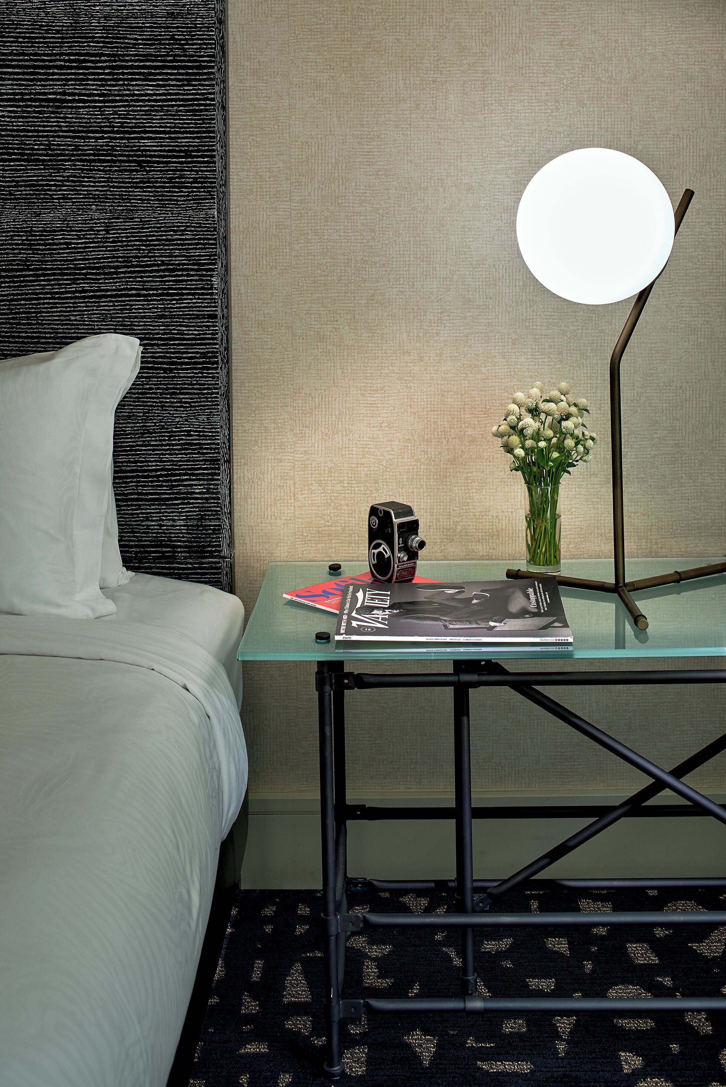 Habitación de hotel con una cama, una mesa de vidrio con una cámara, una lámpara y un florero con flores.