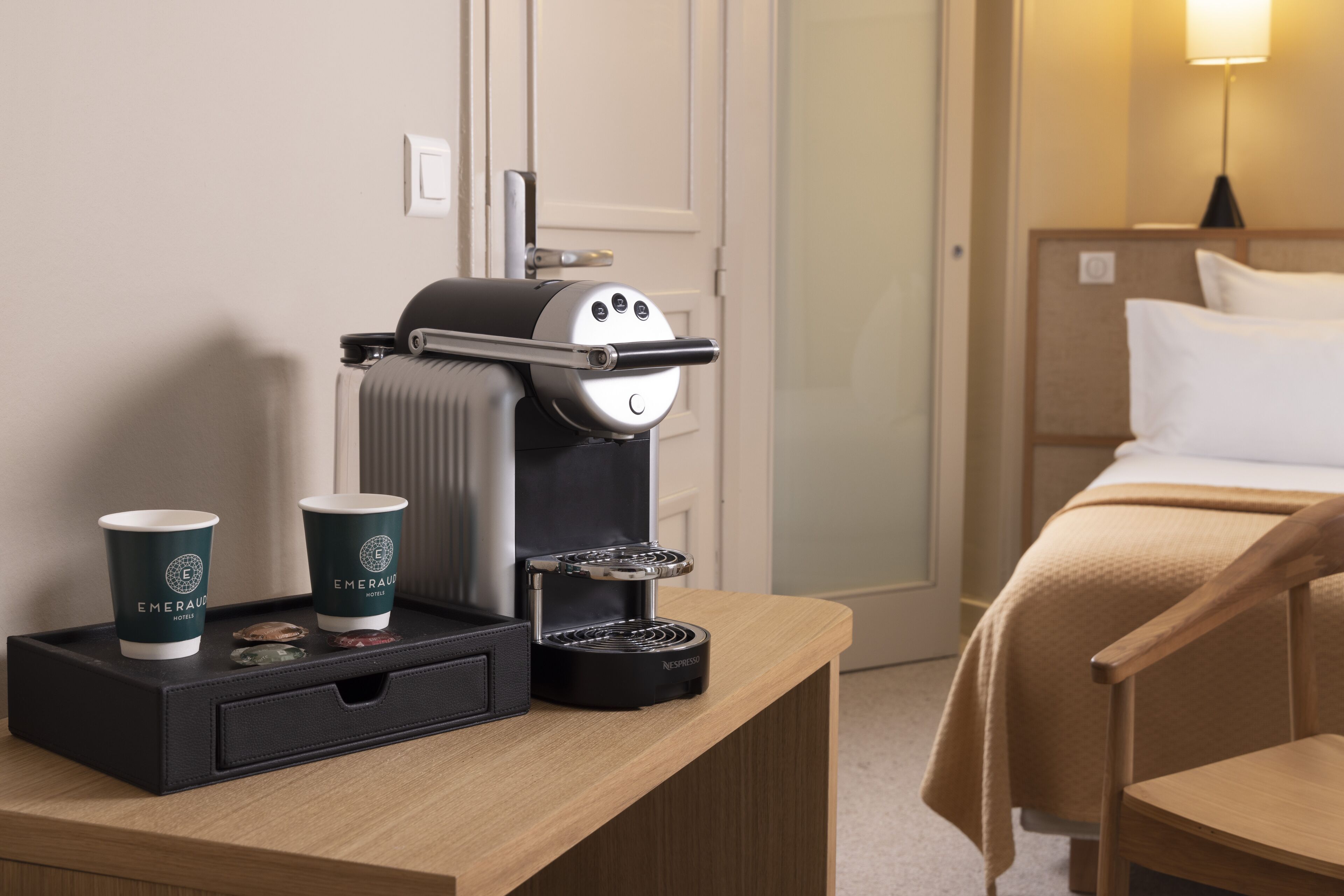 A hotel room with a bed, a wooden nightstand, a coffee maker, and two cups of coffee.