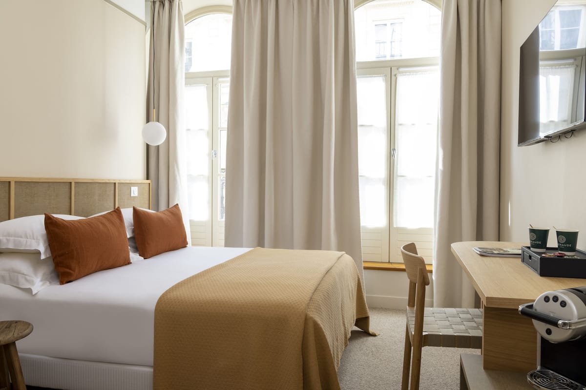 A hotel room with a bed, a desk, and a chair. There is a window with curtains, a television mounted on the wall, and a small table with a lamp.