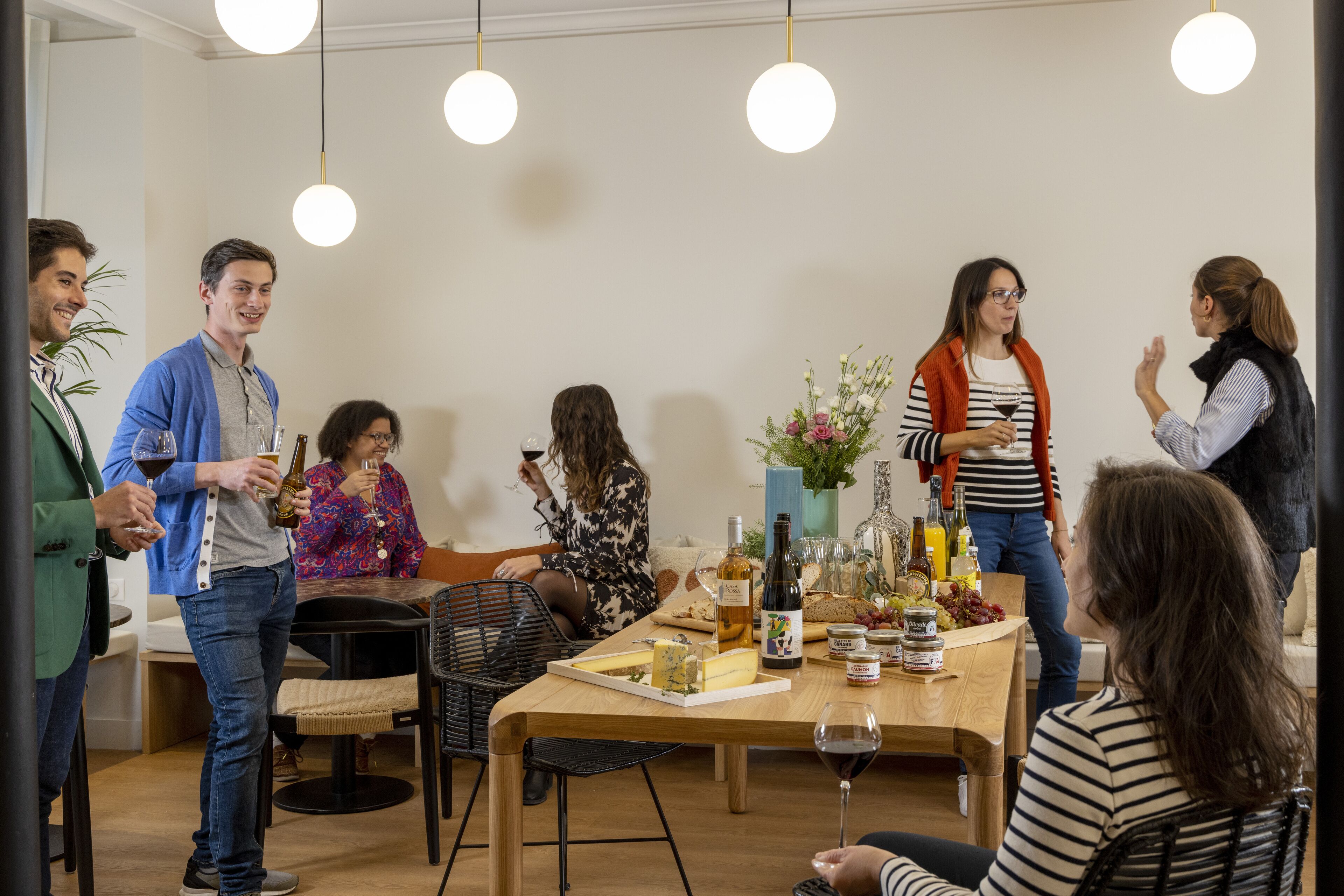 A group of people in a dining area with a table set for a meal, surrounded by chairs and a variety of food and drink items.