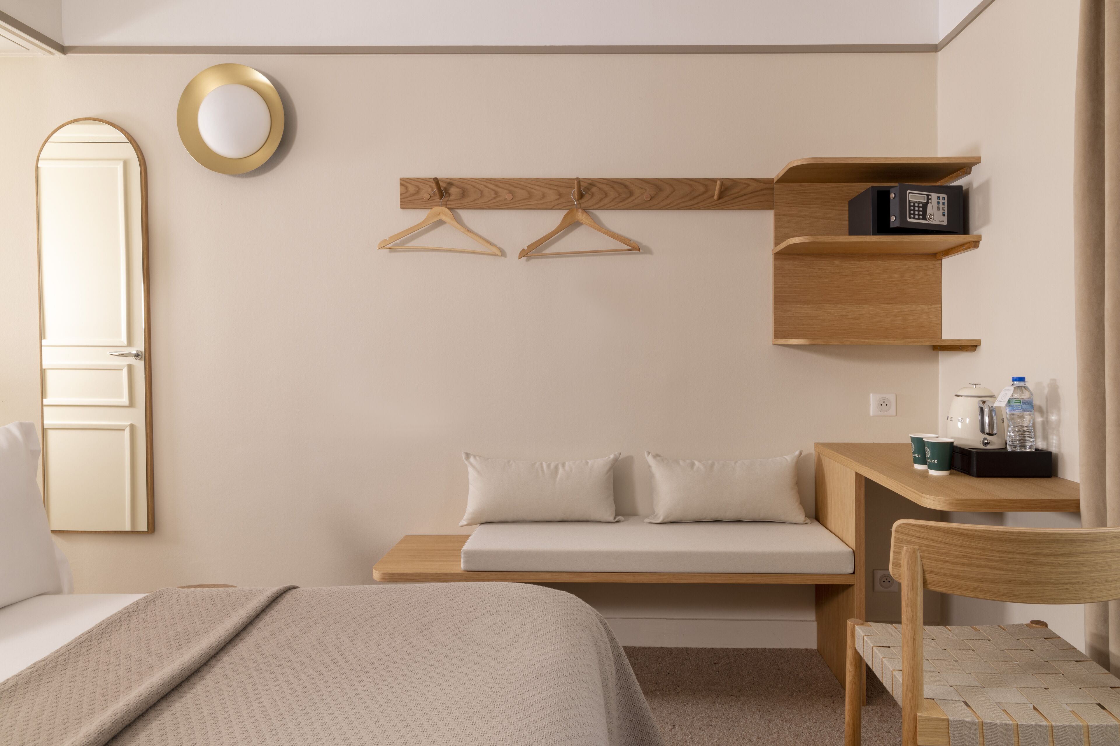 Une chambre d’hôtel comprenant un lit, un canapé, une étagère en bois avec des cintres, un miroir et une petite table avec une bouilloire et une bouteille d’eau.