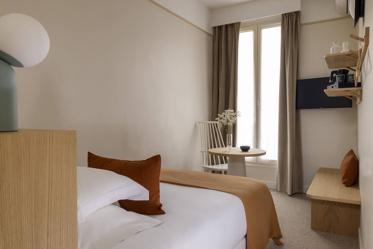 A hotel room with a bed, a small table with a chair, a TV mounted on the wall, and a window with curtains.