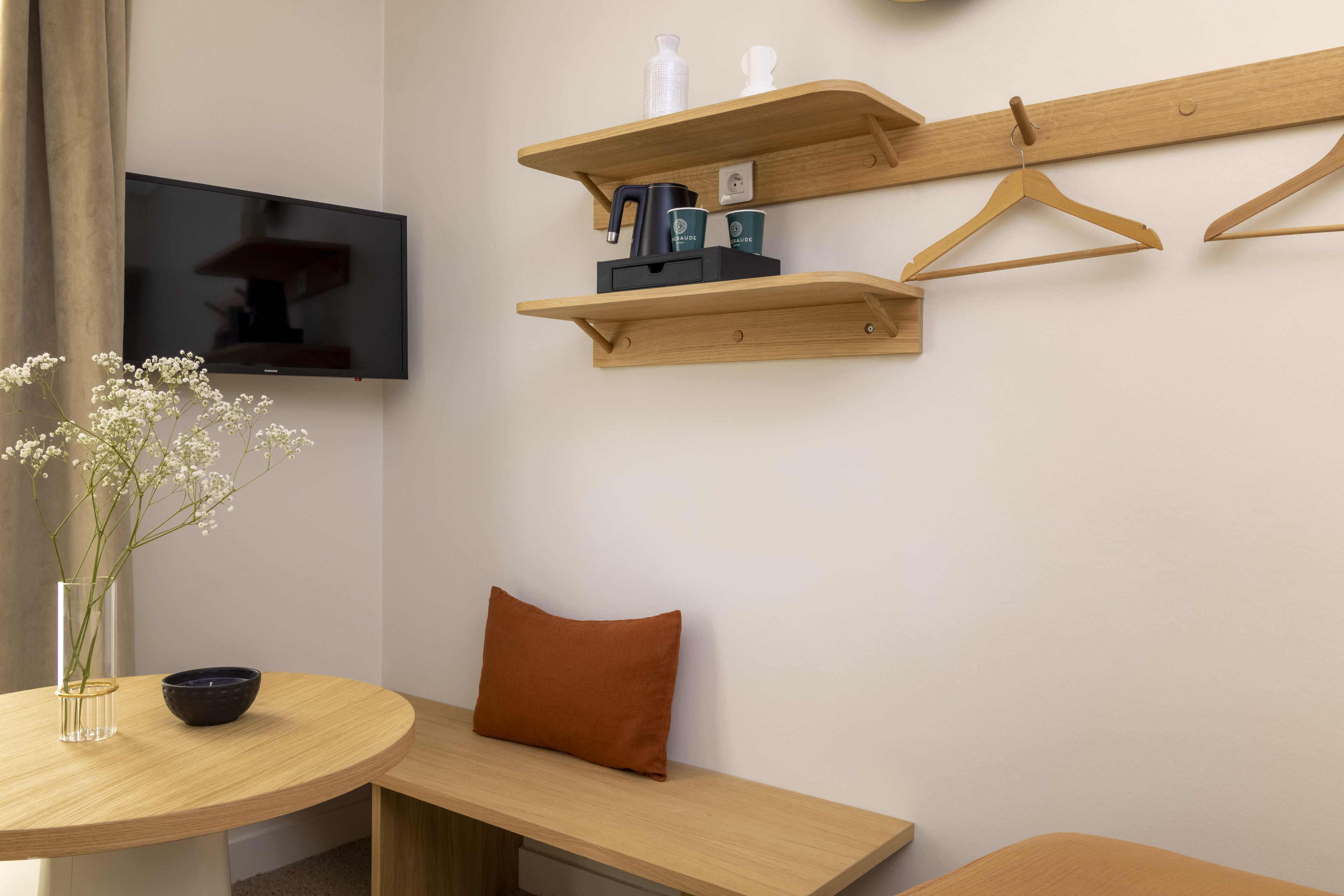 A modern room with a round wooden table, a flat-screen TV mounted on the wall, and wooden shelves with hangers and a kettle.