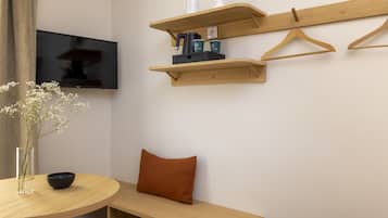A modern room with a round wooden table, a flat-screen TV mounted on the wall, and wooden shelves with hangers and a kettle.