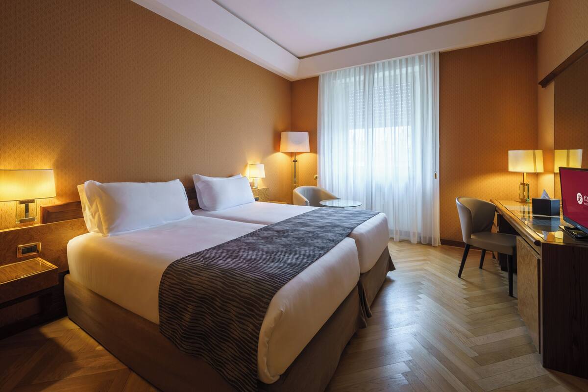 Une chambre d’hôtel avec un grand lit, deux lampes de chevet, un bureau avec une chaise et une grande fenêtre avec des rideaux légers.