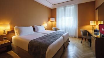Habitación de hotel con una cama grande, dos lámparas de noche, un escritorio con una silla y un ventanal con cortinas translúcidas.