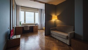 A hotel room with a wooden herringbone floor, a sofa, a desk with a chair, a lamp, and a window with curtains. - Exe Majestic (Naples)