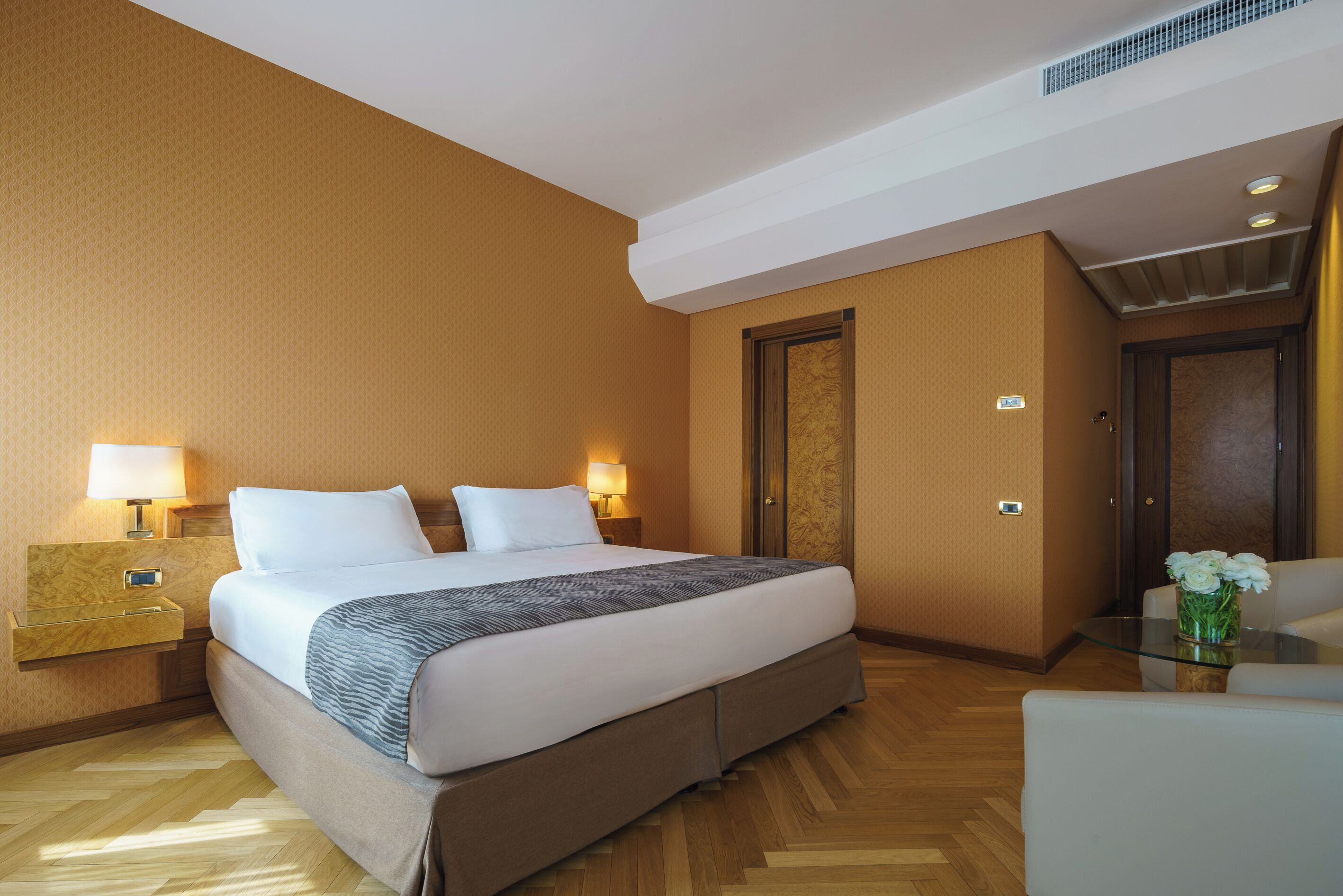 Una camera d'albergo con un letto grande, lampade ai comodini, un pavimento in legno e un tavolino in vetro con un vaso di fiori.