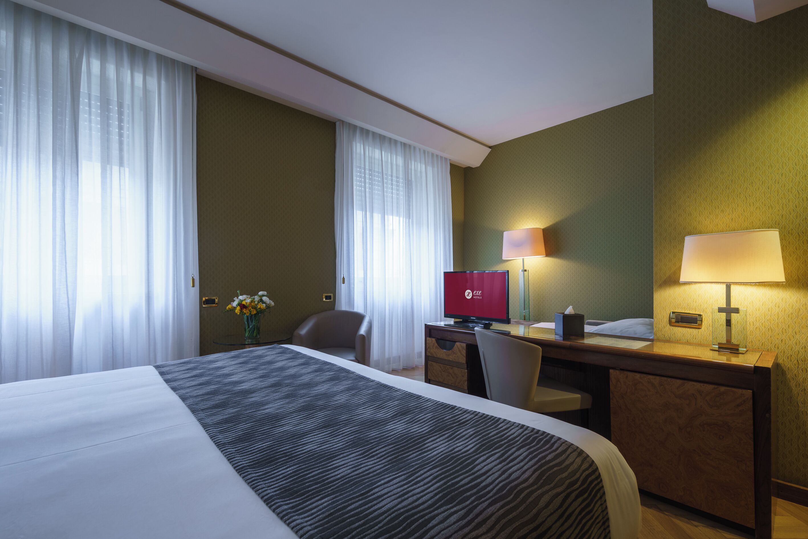 A hotel room with a large bed, a desk, a chair, a television, and a lamp.